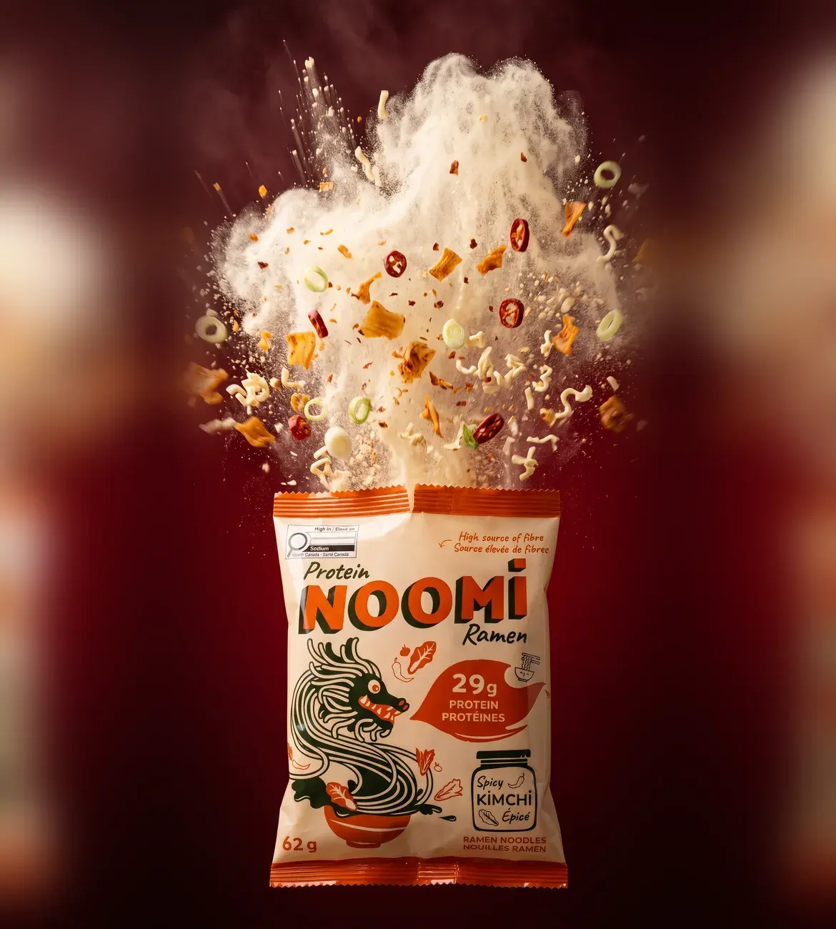 A pack of Noomi ramen bursting open, noodles, kimchi and chilli suspended in the air above it