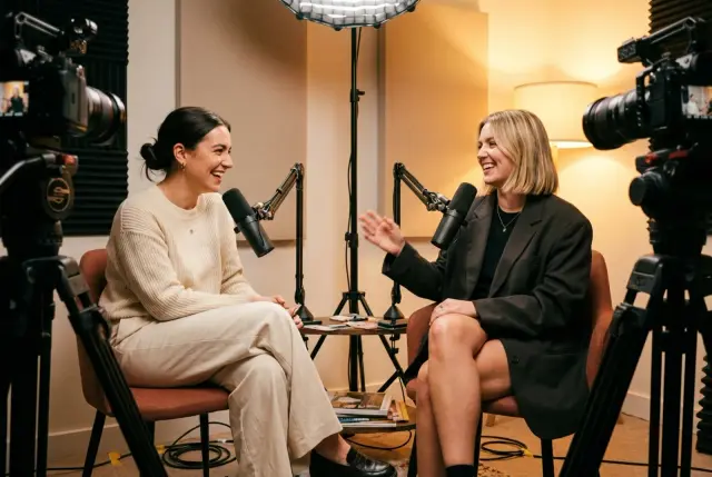 Two women laughing mid-conversation on a podcast set, cameras and microphones around them
